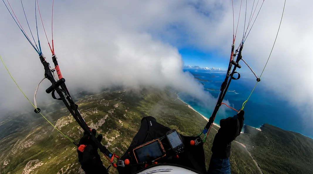 Paraglider with risers visible flying over dramatic coastal cliffs and turquoise ocean